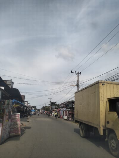 The image depicts a street scene in Kruengmanyang, Indonesia, under an overcast sky. A yellow box truck is prominently featured on the right side of the road, partially obscuring the view of buildings and a sidewalk on the left. These buildings appear to be small shops or market stalls, some with awnings and signage, and a few motorcycles are parked along the side. The road itself is asphalt, and a power pole with numerous wires is visible in the middle ground. Several people are present, some on motorcycles or bicycles, moving along the street. The overall atmosphere is that of a typical urban or semi-urban street in Indonesia.