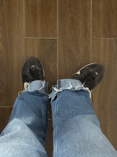 The image shows a pair of feet wearing denim jeans and black Nike sneakers. The jeans are a light blue wash and appear to be rolled up at the ankles, revealing frayed edges. The sneakers are dark with white Nike logos. The floor beneath the feet is a dark brown tiled surface, resembling wood. The scene is captured from a top-down perspective. This image does not contain any information that allows for the determination of a specific location within the city of Masis, Armenia, or any other geographical context.