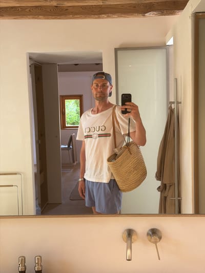 A man is taking a mirror selfie in what appears to be a well-lit room. He is wearing a white t-shirt with a green and red stripe and the "GUCCI" logo, blue shorts, and a dark baseball cap. He is holding a black smartphone in his right hand, angled to capture his reflection. A woven straw bag with leather straps is slung over his left shoulder.

The background shows a reflection of the room, which includes a wooden beam, a closet with clothes hanging, a window with lush green trees visible outside, and what seems to be a metal towel rack or clothing rod. The overall impression is of a casual, possibly vacation setting, given the attire and the straw bag. The lighting suggests daytime. There are two silver door handles or fixtures visible on the wall to the right of the frame. The man's expression is neutral.