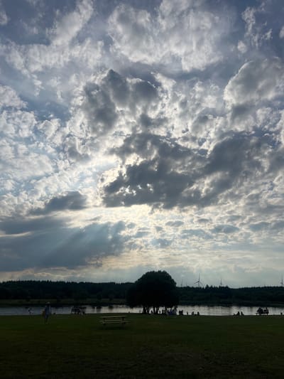 This is a vertical, eye-level, wide shot of a cloudy sky over a body of water with a tree and people present on a grassy area in Ballybought, Ireland. The sky is filled with dramatic, cumulus clouds with rays of sunlight breaking through in the middle ground. The body of water is dark and calm, reflecting the sky. In the foreground, a dark green grassy area has a picnic table and bench. In the midground, along the edge of the water, there is a dark silhouette of a large tree, and to the right of it, a line of wind turbines against the horizon. Several people are scattered on the grassy area and near the water, some are walking, others are sitting. A person in a light-colored outfit and hat walks across the grass toward the water in the middle left of the frame. The scene appears to be a park or recreational area.