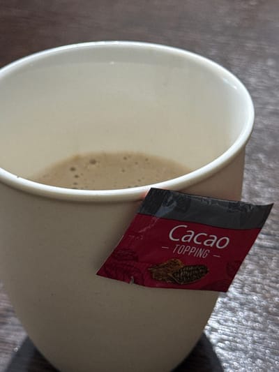 The scene is located in the city of Taperakan, Armenia. A close-up, overhead view depicts a light-colored, off-white ceramic mug, approximately three-quarters full with a light brown, frothy beverage, likely coffee or a similar drink. The surface of the liquid exhibits numerous small bubbles. Resting on the upper exterior side of the mug is a small, rectangular red foil packet with a black top border. The packet is labeled "Cacao - TOPPING" in white sans-serif font, with an illustration resembling cacao beans or pods beneath the text. The mug and the packet are positioned on a dark brown surface displaying a visible wood grain texture, indicative of a table or countertop. No individuals or active interactions are visible in the frame.