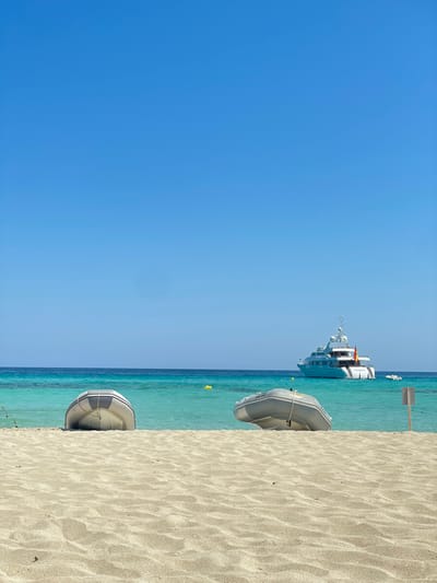 The image depicts a daytime scene on a sandy beach in Formentera, Spain, under a clear, bright blue sky. In the foreground, two grey inflatable tender boats are pulled partially onto the light-colored sand, resting with their bows facing the water. The boat on the left is positioned slightly more to the center, while the one on the right is closer to the right edge of the frame. The sand shows ripples and slight indentations.

Beyond the sand, the water transitions from shallow, clear turquoise near the shore to a deeper blue further out. Several yellow buoys are visible floating in the mid-ground water. A large, white multi-deck motor yacht is anchored in the deeper blue water towards the right side of the frame, approximately mid-distance. A Spanish flag is visible on the yacht's stern. A smaller, white tender boat is positioned behind the larger yacht, to its left. The horizon line is visible in the background where the deep blue water meets the lighter blue sky. No individuals are visible in the scene.