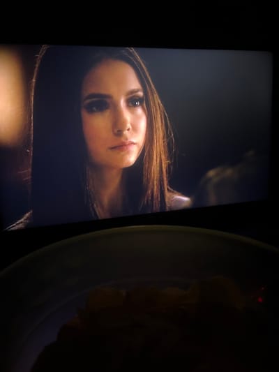 The image shows a close-up of a woman's face on a screen, likely a television or projector, with a bowl of food in the foreground. The woman has long, dark brown hair and is looking off to the side. She is wearing dark eye makeup. The background behind her is dark and out of focus. In the foreground, a shallow bowl contains what appears to be chips or crisps, partially illuminated from above and to the right by a warm light source. The scene is dark overall, suggesting it is evening or a dimly lit room, possibly a home theater setup. There is no visible indication of the location in Каменар, Bulgaria.