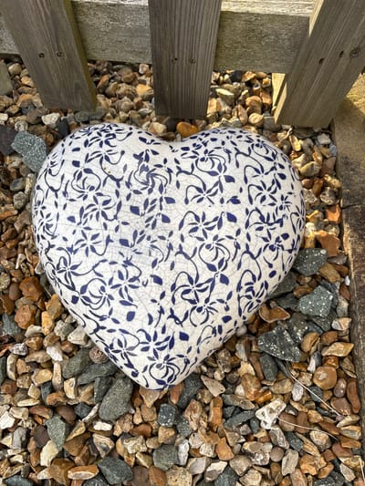 The image depicts an outdoor scene at ground level in Stiperstones, United Kingdom. A heart-shaped ceramic object is centrally positioned, resting on a bed of multicolored gravel. The object is white with a repeating blue floral pattern and features a crackle glaze finish. The gravel consists of irregularly shaped stones in shades of brown, grey, and white. In the upper background, two weathered wooden planks, consistent with fence posts or a garden border, are partially visible, casting shadows onto the gravel. No individuals, animals, or active interactions are present within the frame.