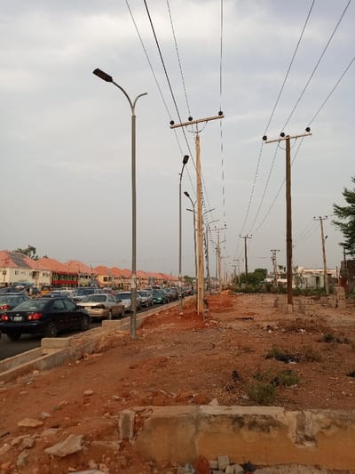 The image shows a street in the city of Way, Nigeria, located at coordinates 9.271 latitude and 12.438 longitude. The street is lined with wooden utility poles, each supporting several power lines. Street lights are mounted on the poles and are facing the street. There are multiple cars driving along the street and parked along the side. The ground is a reddish-brown, likely dirt or clay. There is a curb on the left side of the road, separating it from a sidewalk which is being constructed. The scene is overcast, with some clouds present in the sky. The image was likely taken from a car facing forward as it was driving along the street. There are no pedestrians visible in the scene. Several small trees and shrubs line the opposite side of the street.