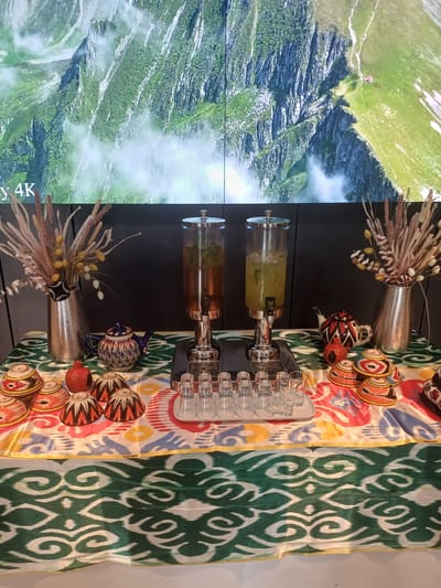 The image displays a table set for a drink service. In the background, a large screen shows a scenic view of green mountains with mist and a small red building on a ridge. In the foreground, two large glass beverage dispensers are positioned side-by-side on a black stand. The left dispenser contains a dark amber liquid, likely tea, and the right dispenser holds a lighter, greenish liquid with ice and mint. To the left of the dispensers, a tall, metallic vase holds a dried floral arrangement. In front of the vase, a small, ornate blue and white teapot sits on a colorful, patterned tablecloth. Next to the teapot, there is a single red pomegranate. A collection of small, decorative bowls with intricate patterns in red, yellow, and black are arranged on the tablecloth. To the right of the dispensers, a tray holds several clear shot glasses. The overall scene suggests a refreshment station, possibly in a hotel or event venue. There are no people visible in the image. The location is identified as the city of Myk, Uzbekistan, implied by the text overlay on the screen, though the image itself does not provide explicit geographical markers beyond that.