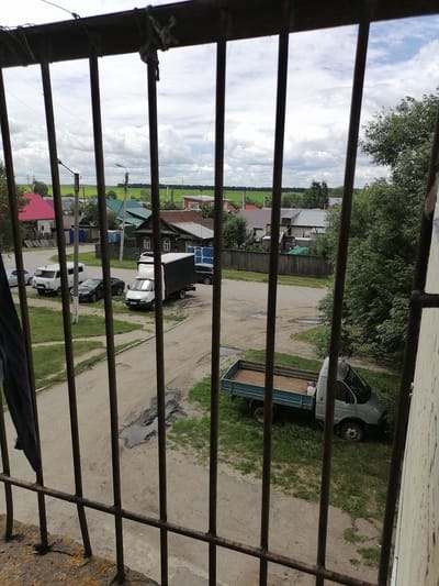 This is a high-angle, full shot taken from behind metal bars, looking out at a residential street scene in the city of Kuznetsk, Russia. The foreground is dominated by the rusty, dark metal bars of a window or balcony railing. Beyond the bars, a dirt and gravel road curves through a village-like setting. On the left side of the road, parked are a white van and a dark sedan. Further down the road on the left, a white box truck is parked. On the right side of the road, a light-colored flatbed truck is parked on a grassy area, partially obscured by trees. The background features several houses with various roof colors, including red, green, and brown. Beyond the houses, there are fields of green crops and a cloudy sky. The overall impression is of a rural or semi-rural residential area.