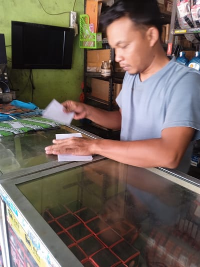 The image shows a man in Kauman, Indonesia, standing behind a counter with a glass display case. He is wearing a light blue t-shirt and is in the act of stacking or sorting white pieces of paper. The counter has stacks of green items wrapped in plastic. The display case contains rows of black boxes with red tops. Behind the man, the background shows a wall-mounted television, shelves with various items, including automotive fluids, tires, and cleaning supplies. Text visible in the image includes product labels like "LUMINOS HID-LED," "MST," "Racing," and "lubricant."
