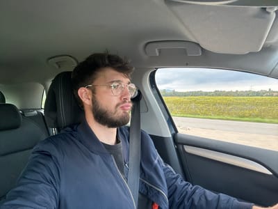 A male individual is depicted in the driver's seat of a vehicle on a road near Châtenois, France. The man, appearing to be in his late twenties or early thirties, has short dark hair, a beard, and a mustache, and wears round, gold-framed eyeglasses. He is dressed in a dark blue bomber jacket over a dark shirt and is secured by a shoulder and lap seatbelt. His lips are pursed, and his head is turned slightly to the right, with his gaze directed out the passenger-side window. The vehicle's interior includes a gray headliner, black seating, and a black door panel featuring a silver accent strip. Visible through the passenger window is an asphalt road with a white shoulder line, bordered by an extensive vineyard with yellowish-green foliage. In the mid-distance, a tree line precedes distant hills under an overcast sky, with a utility pylon faintly visible in the background. The scene suggests the vehicle is in motion.