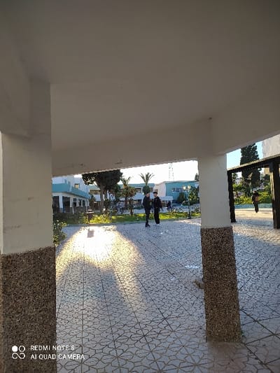 The image captures a perspective from underneath a light-colored overhang, looking out into an open outdoor area.
In the immediate foreground, a wide, plain off-white or light grey wall or ceiling surface occupies the left side and top left corner of the frame, indicating the sheltered viewpoint. On the right side of the image, a vertical pillar or wall section with a rough, speckled, dark grey and brown aggregate texture is prominent, partially framing the view.
The ground area is covered with light-colored tiles, possibly white or cream, adorned with a repeating geometric pattern that resembles interlocking octagons or eight-pointed stars. A striking feature of the scene is a long, bright patch of sunlight that stretches across the tiled ground, extending from the middle of the frame towards the background. This strong, directional light suggests either an early morning or late afternoon time, creating noticeable highlights and shadows.
In the midground, several human figures, approximately three to four individuals, are visible standing or walking on the tiled surface. They appear somewhat indistinct due to distance and being partially in shadow or silhouetted against the brighter light, but they seem to be wearing dark clothing.
Beyond the paved area, the background reveals lush green foliage, including several palm trees, indicating a warm climate. Buildings with light blue or turquoise accents are visible amidst the greenery, suggesting an institutional, residential, or resort-like setting. The sky above is bright and clear.
In the bottom right corner of the image, a white watermark is present, displaying the text "REDMI NOTE 8" and "AI QUAD CAMERA", accompanied by two small circular logos, indicating the device used to capture the photo.