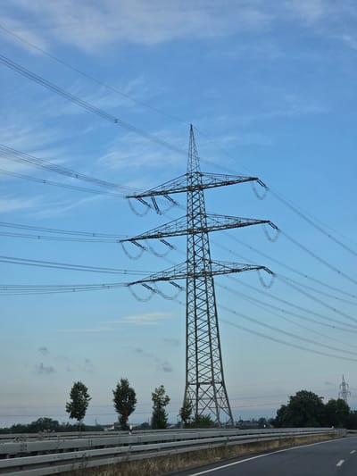 The image shows a large electricity pylon and high-voltage power lines against a blue sky with scattered wispy clouds. In the foreground, a road with a guardrail is visible. Several trees are planted along the side of the road, with more trees and some distant industrial structures in the background. The scene appears to be located on the outskirts of Rüsselsheim am Main, Germany, likely near a transportation route given the presence of the road and guardrail. There are no people or active interactions depicted. The environment suggests a semi-industrial or rural setting with infrastructure for power transmission.