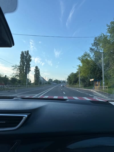 The image depicts a view from inside a vehicle looking out at a road in Vatra, Moldova. The sky is a clear blue with wispy clouds. On the left side of the road, there are tall trees and power lines. On the right side, there is a bus stop with a blue shelter and a yellow railing. The road itself has white lines marking the lanes and a crosswalk with red and white stripes in the foreground. There is one car visible driving in the distance. The overall scene appears to be a suburban or semi-rural area during daylight.