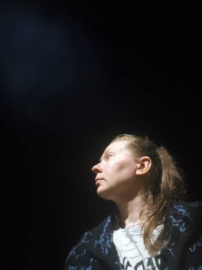 A close-up, side-profile medium shot captures a woman with blonde hair pulled back in a ponytail, looking upwards and to the left in what appears to be a dark, possibly indoor, setting. She is wearing a dark jacket with a blue, abstract pattern over a white t-shirt with some writing on it. A spotlight or light source illuminates her face from the top left, creating highlights and shadows. The background is predominantly black, with a subtle gradient of light near the top left. The woman's skin is fair, and she has a prominent nose and ear. The image is framed such that the woman's face occupies the lower right quadrant of the frame, with the majority of the image dedicated to the dark background. The location of Il'mino, Russia, is not depicted or suggested in this image.