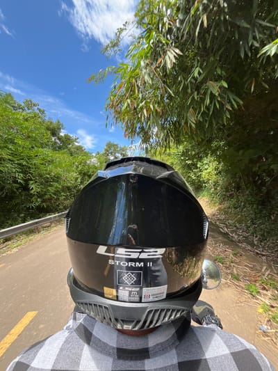 In a first-person perspective, a black LS2 Storm II motorcycle helmet is the central focus of the image. The helmet is adorned with stickers, including one with the text "TEM CHONG HANG GIA" and another displaying certification marks like "ECE 22.06" and "1500 g". The rider, wearing a gray and white plaid shirt, is on a motorcycle. They are traveling on a paved road with a yellow line marking, bordered by lush green foliage and bamboo trees. The scene appears to be in a rural or natural setting, likely Son Dien, Vietnam, given the context. The sky is a vibrant blue with scattered white clouds, suggesting good weather. The road curves gently to the right, disappearing into the verdant landscape.