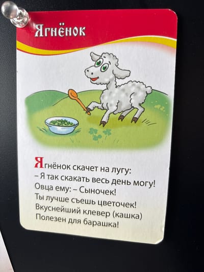 A laminated card featuring a cartoon drawing of a lamb is attached to a dark surface, likely a wall or bulletin board, by a silver pushpin in the upper left corner. The card has a red border at the top with the Russian word "Ягнёнок" (Lamb) written in white stylized font. Below this, a yellow wavy line separates the red border from the white card's main illustration. The illustration depicts a fluffy white lamb with green eyes, holding an orange spoon and standing on a green hill next to a bowl of green food. The text below the illustration is in Russian and appears to be a poem or story about the lamb. The background of the image is a dark, textured surface.