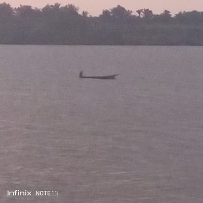 The photograph depicts a flat body of water, presumably a river or large lake, reflecting a muted, overcast sky. In the middle distance, a small wooden boat is visible, with a person in the boat, likely navigating the water. The shoreline, densely vegetated with trees, is faintly discernible in the background under a sky that hints at either dawn or dusk. The photograph was taken in Atani, Nigeria.
