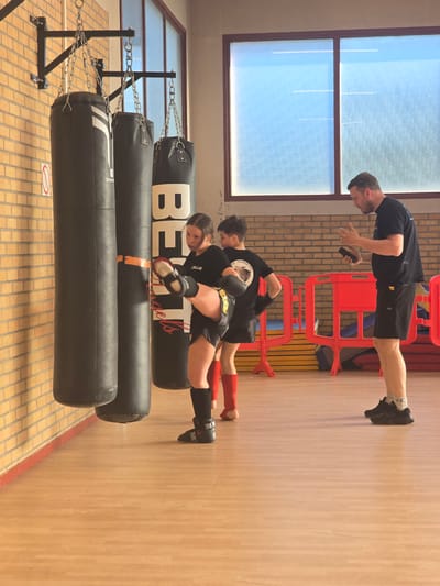This media file captures an indoor training session at a combat sports facility, likely a gym or sports hall, in Leers, France. The scene features three individuals: an adult male supervising two younger trainees.
The adult male, with a beard and dark hair, stands upright in a black t-shirt and shorts, observing the trainees below him. He holds what appears to be a phone or remote control in his hand. The two younger trainees are engaged in an exercise, positioned horizontally in a plank-like pose atop black heavy punching bags. Both are dressed in black t-shirts and shorts, with protective shin guards and boxing gloves/hand wraps. One trainee wears red shin guards and black gloves, while the other has black shin guards and black gloves with yellow details. The latter also has the name "CAILLIE" on their t-shirt and "METAL" on their hand wrap. The other trainee's shirt has "DESORDRE" visible.
In the foreground, three black punching bags of varying sizes are suspended by chains from a robust black metal frame. The top bag prominently displays "BELT" and "Angels", while another shows "DOMYOS". The facility has a light wooden floor, and the walls are a combination of light-colored brick and plain sections. Large windows with textured glass allow ample natural light into the space, indicating it is daytime. Red plastic barriers are also visible in the background, and a small, red warning sign is on the floor. The overall impression is one of focused training and coaching.