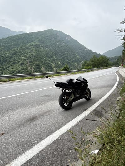 A black motorcycle is parked on the right shoulder of a winding asphalt road, set against a backdrop of densely forested mountains under an overcast sky. The motorcycle, a sleek sport or street model, faces away from the camera, positioned on a gentle curve of the road. White road markings, including a solid line along the shoulder and dashed lines further ahead, are visible on the gray pavement.

To the left of the road, a metal guardrail runs parallel to a steep, tree-covered slope that descends into a valley. Towering mountains, thick with green foliage, dominate the background, their upper sections blending with the muted gray of the cloudy sky. In the distance, a red and white chevron road sign is visible, indicating a curve or hazard. The right side of the road has sparse vegetation and curb detailing.

The scene suggests a stop during a journey through a scenic, mountainous region, potentially near Krichim, Bulgaria. The weather is cloudy and calm, with no direct sunlight. No people are present, and no legible text can be identified on any objects or signs.