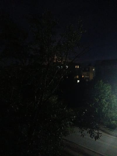 A nighttime view of an apartment building and surrounding trees is captured in this photograph, likely taken in Kuznetsk, Russia. The building, illuminated by interior lights in several windows, appears to be multi-story. Dense foliage from trees partially obscures the view of the building and the paved area in the foreground, which could be a road or pathway. The overall scene is dark, with the limited light sources creating a sense of depth and mystery. The image conveys a quiet, possibly urban, environment at night.
