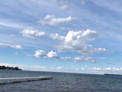 The image shows a body of water, likely a lake, with a white wooden dock extending into it from the left. The water is a deep blue-gray and appears slightly choppy. The sky above is a bright blue with scattered white cumulus clouds. In the distance, on the left side of the frame, there is a shoreline with trees and a few buildings. On the right side of the frame, there is a distant island or landmass with trees. The scene evokes a sense of calm and nature. The location is Innisfil, Canada, suggesting it is likely Lake Simcoe. There are no people visible in the image.