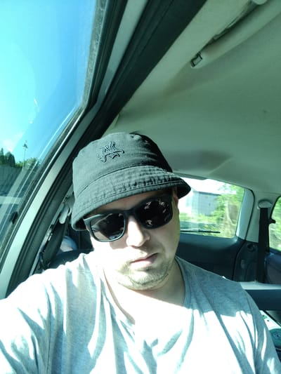 In this selfie taken from inside a car, a person is seen wearing a black bucket hat with an emblem and black sunglasses. The person has a light beard and is wearing a light grey t-shirt. The car's interior is visible, including the headliner and a portion of the window. Sunlight streams in, creating strong shadows on the person's t-shirt. Outside the car window, trees and buildings are visible, suggesting an outdoor setting, possibly in the city of Tehov, Czechia.