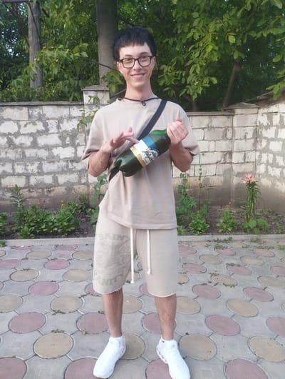 A young man with glasses and black hair is standing outdoors, holding a green bottle. He is wearing a light-colored t-shirt and shorts, and white sneakers. The background consists of a brick wall and trees. The ground is paved with circular stones. The man is smiling. He appears to be in the city of Fălești, Moldova, based on the context provided.