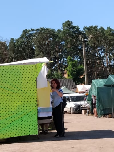 In Irshansk, Ukraine, a woman with red hair is examining items displayed under a bright green canopy patterned with small dark shapes. She is wearing a light blue shirt and black pants. To her left, hanging clothes are visible, including a white t-shirt and a yellow garment. In the background, a white van is parked, and several green tents are set up. The scene appears to be an outdoor market under a clear blue sky, with a line of pine trees bordering the area.