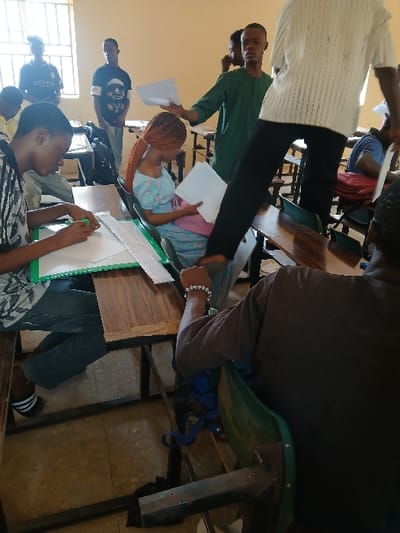 The image displays an indoor scene within a building in Ugi Majowel, Nigeria, resembling a classroom or administrative hall. The area is occupied by approximately eight individuals, predominantly young adults, engaged in activities involving documents. In the foreground-left, a young male is seated at a wooden desk, focused on writing or marking a sheet of white paper with a green marker, a green folder positioned to his left. To his right, the back of a person, likely male, is visible, seated in a green plastic chair, wearing a dark brown shirt and a white beaded bracelet on their left wrist. In the mid-ground, a young female with reddish-brown braided hair, wearing a light blue patterned off-the-shoulder dress, is seated at a desk, holding and reviewing white papers. A male in a dark green traditional garment stands nearby, holding multiple white documents, appearing to distribute or collect them. Another individual, partially visible in the mid-ground, wearing a white striped shirt and dark trousers, is depicted in motion, stepping over a desk. In the background, near a window fitted with vertical bars, two other males are standing, one wearing a dark t-shirt with "ETIHAD" visible text. The room features light-colored walls, a tiled floor, and multiple wooden desks with black metal frames paired with green plastic chairs, indicating a structured environment.