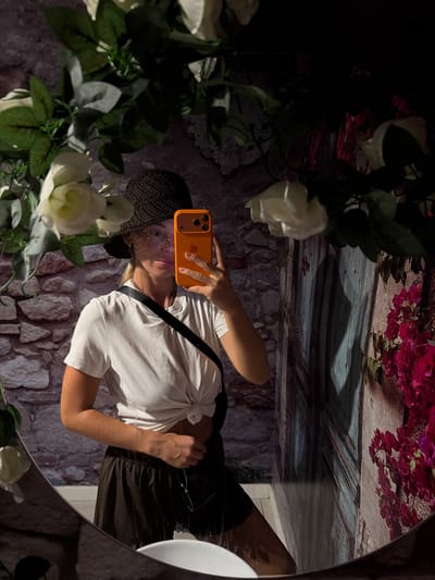 In Urbanització Cales I Talaies, Spain, a woman wearing a black sun hat and a white knotted t-shirt is reflected in a mirror. She is holding an orange smartphone, taking a selfie. She is also wearing dark shorts and a black strap is visible across her chest. The mirror reflects a backdrop of a stone wall, a rustic wooden door, and artificial white roses and pink bougainvillea.