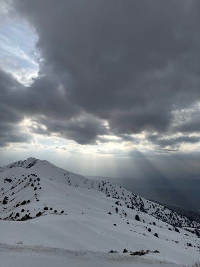 The image displays a snow-covered mountain landscape in the Nurekata region of Uzbekistan. The sky is largely overcast with extensive dark grey clouds, from which multiple distinct rays of sunlight penetrate downwards towards the distant horizon. Snow blankets the prominent mountain ridges and slopes visible across the midground. Sparse, dark bushes or small evergreen trees are scattered across the snow-laden terrain. In the lower mid-ground, a series of small, dark, dome-shaped structures or shelters are arranged in a horizontal line on a gentle slope. The immediate foreground reveals a snow-lined edge, consistent with a road or track. No individuals, animals, or active interactions are present. The environment appears cold and mountainous, indicative of winter or early spring conditions.