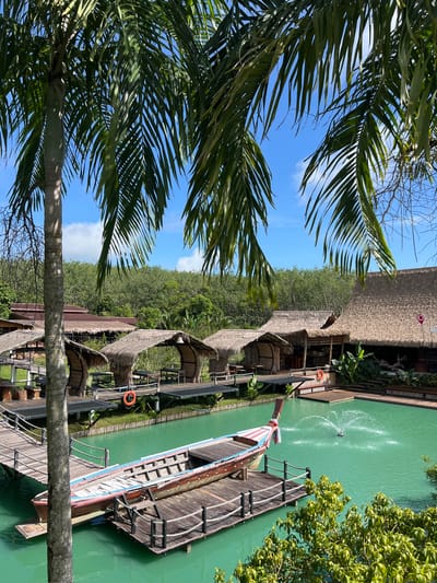 This media file depicts a scenic waterside resort or restaurant in Tambon Pa Kae Bo Hin, Thailand. The foreground is dominated by the lush green fronds of a palm tree, framing a view of the tropical setting. The scene is bathed in bright sunlight, suggesting it's daytime.
The central focus is a vibrant turquoise body of water, likely a river or canal, where a long, narrow wooden boat with a red, white, and blue striped trim is docked. The boat has a traditional design and appears to be a prominent feature of the location. In the water, a fountain or water jet creates a dynamic spray.
Along the water's edge, a series of thatched-roof structures, possibly huts or dining areas, are connected by wooden walkways. These structures have a rustic, natural aesthetic. Orange life preservers are visible, indicating a focus on water safety. Beyond the resort area, a dense forest of green trees stretches towards the horizon under a clear blue sky with scattered white clouds.
The overall impression is one of tranquility and natural beauty, characteristic of a tropical vacation destination. No people are explicitly visible engaging in activities, but the presence of dining areas and the boat suggests leisure and potential tourism. The time of day is clearly daylight, and the weather appears to be sunny and pleasant. The location cues point to a tropical environment, likely Southeast Asia, specifically Thailand as indicated by the location context.