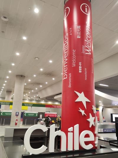 The image captures a scene inside an airport, specifically at Arturo Merino Benítez International Airport located in Pudahuel, Chile. A large, red cylindrical column serves as a welcome sign, featuring the word "Welcome" and its translations in multiple languages like Japanese, Korean, Spanish, and French. The column is decorated with white stars. In the foreground, a white three-dimensional sign spells out "Chile."

In the background, there's an area with "Nothing to Declare" signs in both Spanish and English, along with customs checkpoints. Passengers are moving through the airport, wearing backpacks and outerwear. The environment is well-lit with numerous overhead lights. A baggage claim area is visible in the right background, alongside a sign for "Mobility Assistance." The airport's interior exhibits modern design, and a fire extinguisher is mounted on a pillar.