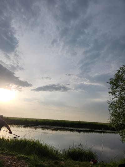 The image depicts a serene scene of a person fishing on the bank of a calm body of water, likely a river or lake, in Atameken, Kazakhstan. The sun is low in the sky, casting a warm, golden light that suggests either early morning or late evening. The sky is partly cloudy, with a mix of dark and light clouds creating a dynamic backdrop. Lush green grass and reeds line the bank of the water, and the water itself reflects the sky and surrounding vegetation, creating a mirror-like effect. A fishing rod is held by the person, who is partially visible in the foreground, suggesting an active fishing session. The overall atmosphere is peaceful and tranquil, evoking a sense of quiet contemplation and connection with nature. There is no visible text in the image.