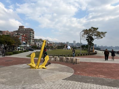 A public space features a large yellow anchor statue in the foreground, resting on a concrete base. Beyond the anchor lies a grassy area with a few scattered figures, some trees, and the word "TSUNGSO" constructed from individual concrete blocks. A paved path with people walking along it runs parallel to the river in the background. Buildings and a city skyline are visible in the distance under a partly cloudy sky.