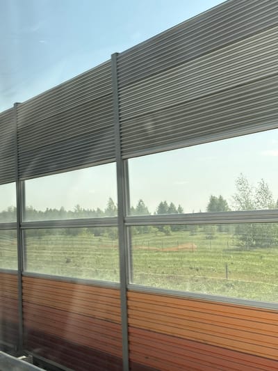 The image shows a partial view of a structure, possibly a bus stop or a covered walkway, featuring a corrugated metal roof on the left and a wooden slat roof on the right. Between the two roof sections are glass windows offering a view of a green, grassy field with trees. The scene is slightly overexposed, obscuring detail in the landscape. No people or other subjects are visible. It is impossible to confirm the location of the city of Сапуголи, Russia from this image.