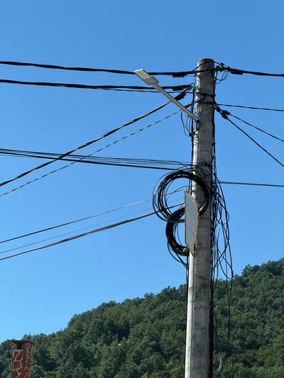 The image shows a utility pole in Beli Osam, Bulgaria, against a clear blue sky. The concrete pole is heavily laden with numerous black electrical and communication wires, some of which are coiled around the pole. A street lamp is attached to the upper section of the pole. In the background, a lush green, tree-covered hillside is visible, suggesting a natural or semi-rural setting. A portion of a decorative chimney, with a textured surface, is present in the lower left corner of the frame. There are no people or animals visible in the image.