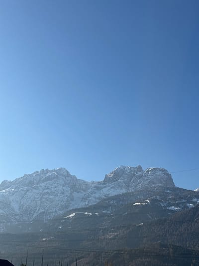 The image depicts a winter mountain landscape in Nußdorf, Austria, under a clear, cloudless blue sky. Snow-covered, jagged mountain peaks dominate the background, appearing hazy at their base. The midground consists of densely forested slopes, primarily with coniferous trees, interspersed with patches of snow. Across the lower portion of the frame, several parallel power lines traverse horizontally. A dark, indistinct structure, possibly a roof or building edge, is visible in the bottom left corner. The scene is illuminated by bright sunlight, casting subtle shadows on the mountain faces. No people, animals, or specific actions are visible.