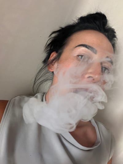 A close-up, slightly downward-angled, self-portrait captures a person's face partially obscured by a cloud of white vapor. The individual has dark hair pulled up loosely, and appears to be exhaling. Their eyes are open and looking towards the camera, with visible eyebrows and some freckles on their cheeks. They are wearing a light gray top. The background is a plain, light-colored wall, with a hint of a brown surface visible at the bottom left. The image does not provide any information about the location in Koltushi, Russia.