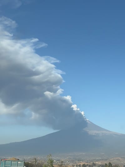A towering mountain shrouded in smoke dominates the right side. Wispy clouds extend into a clear blue sky to the left. A town nestles at the base of the mountain, with foliage visible in the foreground. The photo has been rotated to a vertical orientation.