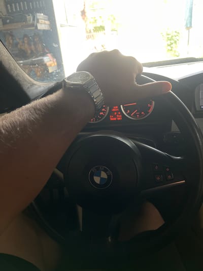 The image provides a first-person perspective from inside a BMW car, showing a person's right arm and hand holding the steering wheel. A prominent feature is a metallic watch with a complex, possibly chronograph, dial and a metal bracelet on the wrist. The car's interior is visible, including the black multi-function steering wheel with the iconic BMW logo, and control buttons for media, phone, and custom functions.
The instrument cluster, illuminated with a warm red-orange glow, is visible through the steering wheel spokes. It displays key information: the car is in "P" (Park), the speed is "0 km/h", and the ambient temperature is "+22.5°C". The time "9:30" and odometer readings "315192" and "325.1" are also visible.
Bright sunlight streams into the car from the left, illuminating the hand and arm, and creating reflections on the side window. These reflections reveal what appears to be a cluttered garage or workshop environment, with various objects, tools, and potentially shelving units. The strong light suggests it is daytime, and the car is likely stationary, parked in a garage with its door or entrance open to the outdoors in Troyanovo, Bulgaria. The driver's thumb rests on the top-left portion of the steering wheel.