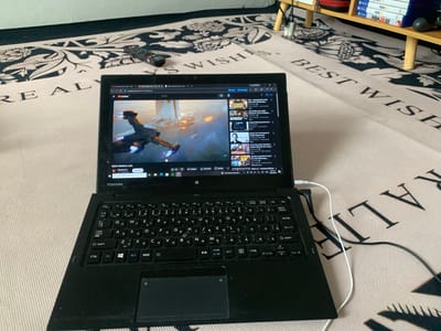 The image shows a black Toshiba laptop open on a patterned rug. The screen displays a YouTube video with a thumbnail of a futuristic aircraft scene, alongside a YouTube sidebar with various video recommendations in Indonesian. Above the laptop, on the rug, lies a black remote control. In the background, a wooden shelf holds stacked game boxes including "FIFA 16", "FIFA 15", and "NBA2K18". Cables are connected to the laptop. The rug has a light color base with darker ornate designs and the phrase "BEST WISHES". The location appears to be indoors. There is no information to indicate this is in Blangbladeh, Indonesia.