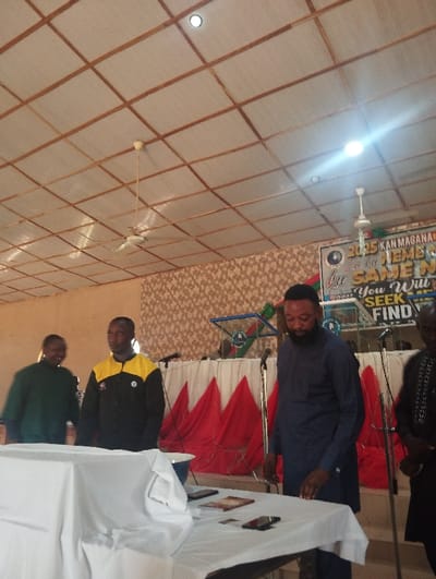 This image captures an indoor event, likely a community gathering or religious service, in Zonkwa, Nigeria. Four adult males are visible. In the immediate foreground, a white-draped table holds a white bowl, two smartphones (one black, one red-brown), and several papers or small books. A bearded man in a dark blue, long-sleeved tunic stands to the right of the table, looking down at its contents with his right hand near the surface. To his left, another man wears a black and yellow collared long-sleeved shirt. Further left, a third man is partially visible, wearing a dark green top. On the far right, a fourth man is partially visible, dressed in a black garment with white embroidery.
Behind the table, four microphones on stands are positioned in front of a stage area. The stage backdrop consists of a brown and tan patterned wall, partially obscured by red pleated fabric draped below a white border. A banner mounted above the stage reads "2025 KAN MAGANA" and "You Will SEEK FIND," with other text partially visible. A green and blue structural element, possibly an emblem, is also present on the stage. The ceiling is composed of light-colored rectangular panels supported by a grid of wooden beams, illuminated by recessed lights and featuring two hanging ceiling fans.