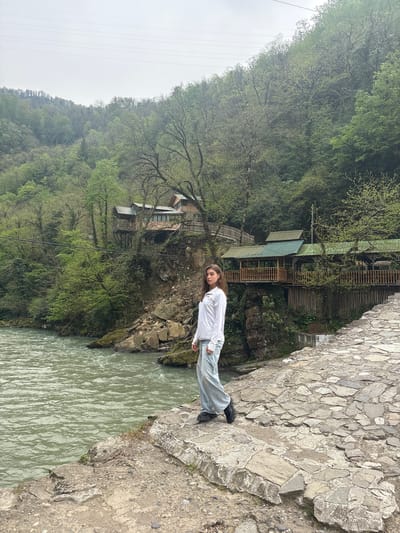 A young woman stands on a stone bridge overlooking a river in Kveda Makhuntseti, Georgia. She is dressed in a white shirt and light-colored pants, with dark shoes. The bridge is constructed of irregular, flat stones. Below, a river flows, and on the opposite bank, a lush, green hillside rises. Wooden structures, possibly homes or shops, are nestled among the trees. The sky is overcast, giving the scene a muted, natural light.