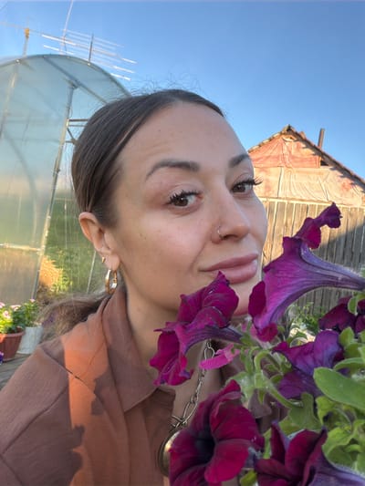 The image is an over-the-shoulder, selfie-style shot of a young woman with dark hair and brown eyes. She is wearing a brown top and a silver chain necklace. Her face is tilted upwards, with purple petunias framing her face. She has a nose ring and wears gold hoop earrings. The background shows a rustic wooden building with a corrugated roof, a greenhouse, and a clear blue sky, suggesting an outdoor setting. The lighting indicates it is daytime, likely late afternoon or early evening, with the warm glow of the sun casting shadows on her clothing and the building. The overall mood is relaxed and natural. The image does not contain any visible text.