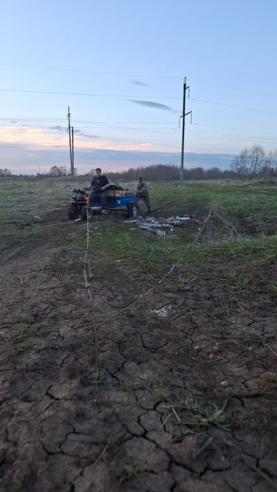 Two men are next to a small, blue four-wheeled vehicle in a field. One man is standing near the vehicle, and the other is on it. There are some pieces of material or debris on the ground near the vehicle. A young tree sits left of the vehicle, and the ground is cracked. Power lines stretch across the sky. The sky is light blue near the horizon with some pink and orange hues. The location is Dubrova, Russia.