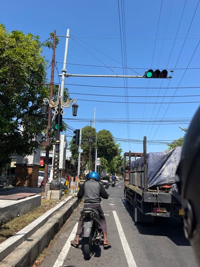 In Pagerwojo, Indonesia, a street scene unfolds under a clear blue sky. The image is captured from the perspective of someone on a motorcycle, looking forward. Ahead, a person wearing a blue helmet and a grey jacket rides a motorcycle. To the right, a large truck with a blue tarp covering its cargo is visible, driving in the same direction. The road is bordered by a concrete curb and sidewalk, with trees and buildings lining the street. A traffic light hangs over the intersection, showing a green light. Decorative street lamps are also present, adding to the urban landscape. The overall impression is one of a typical day on a busy street in Indonesia.
