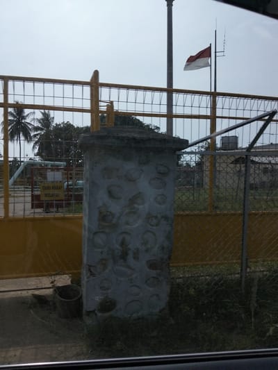 This image depicts an outdoor scene in Blang Mee, Indonesia, focused on a gate and surrounding industrial or utility area. A large, yellow fence with a diamond mesh pattern encloses the property. In the foreground, a weathered concrete pillar, textured with circular indentations, stands to the right. Behind the pillar and fence, a red gate is visible with a yellow sign that reads "TAMU HARAP MELAPOR," meaning "Visitors Please Report." Beyond the gate, pipes and industrial equipment are present, suggesting a functional facility. A red and white Indonesian flag flies on a pole in the background, indicating national presence. Coconut palm trees are visible on the left, typical of the region. The sky is overcast. The scene appears quiet and not actively populated.