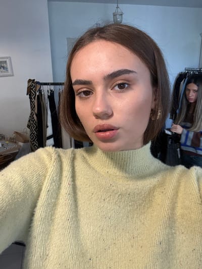 The image depicts an indoor scene, likely a retail clothing boutique or a dressing area, located in Kfar Yona, Israel. In the foreground, a young adult female faces the camera directly, appearing to take a self-portrait. She has shoulder-length brown hair, prominent dark eyebrows, and is wearing a light yellow, high-necked, fuzzy-textured sweater speckled with small colorful fibers. Her lips are slightly parted and appear to have a glossy finish. In the background, to the right of the primary subject, a second female individual is partially visible and out of focus. She has longer, lighter brown hair and is wearing a horizontally striped sweater. Her facial expression is difficult to ascertain but appears neutral to slightly unamused. The environment includes dark-colored clothing racks displaying various garments to the left and right, a light-colored wall, a small framed picture with a light mat on the upper left wall, and a light-colored pendant light fixture suspended from the ceiling. A surface to the left holds several small objects and a brown paper bag.