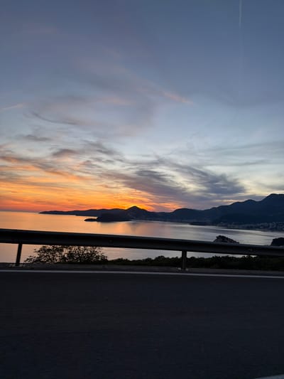 The image captures a scenic view of a coastal landscape at sunset, likely in the vicinity of Blizikuće, Montenegro. The foreground features a dark asphalt road and a metallic guardrail, suggesting the perspective is from a road overlooking the sea. Beyond the guardrail, a body of calm water, likely the Adriatic Sea, stretches towards the horizon. The water reflects the warm hues of the setting sun.
A rugged coastline with dark, silhouetted mountains forms the backdrop. Nestled along the coast is a town or settlement, with buildings faintly visible against the darkening landscape. A small island or rocky outcrop is present in the water, adding to the picturesque setting.
The sky above is a dramatic canvas of color. The left side of the sky is dominated by vibrant orange and yellow streaks, indicative of the setting sun. These warm colors gradually transition to softer pinks and purples, and then to a muted blue-grey in the upper portion of the sky. Wispy clouds are scattered across the sky, catching the last rays of sunlight. A faint contrail is visible in the upper right section of the sky.
The scene is devoid of people or any discernible action, presenting a static, natural landscape illuminated by the twilight. The overall impression is one of tranquility and natural beauty during the transitional period between day and night.