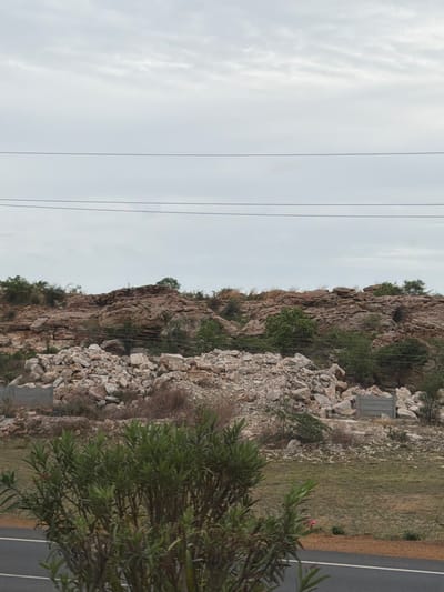 An outdoor, medium shot captures a rocky hill with a pile of stones in the foreground. The sky is overcast with faint clouds. Three parallel power lines stretch horizontally across the upper third of the frame. The hill is covered with brown and grey rocks, with sparse green vegetation and trees dotting its surface. In the lower part of the image, a large heap of light-colored stones and debris is visible. To the left of the stones, a low concrete structure is partially visible. A patch of dry grass and some green shrubs are in front of the pile of stones. A vibrant green bush with narrow leaves occupies the bottom left corner. A paved road runs along the bottom edge of the frame. The scene depicts a rocky, undeveloped area with signs of construction or demolition.